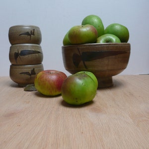 Könnte beinhalten: Eine braune Keramikschüssel gefüllt mit grünen Äpfeln steht auf einem Holztisch neben einem Stapel von drei braunen Keramikbechern.