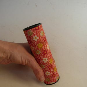 May include: A cylindrical kaleidoscope with a floral pattern in red, white, yellow, and green. The kaleidoscope has a black rim at both ends. The object is held in a hand.