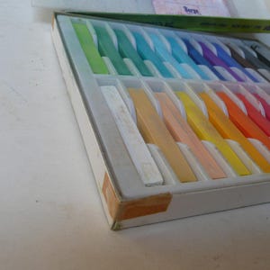 May include: A box of soft pastels in a variety of colors, including green, blue, purple, yellow, orange, and red. The pastels are arranged in a white tray with individual compartments. The box is white with a green lid.