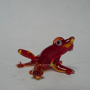 Puede incluir: Una pequeña figura de rana de vidrio rojo con ojos blancos. La rana está sentada sobre una superficie blanca.