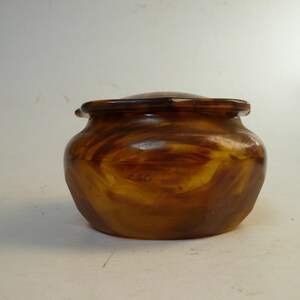 May include: A small, round, lidded container crafted from a translucent, amber-colored material with a swirling, marbled pattern of dark brown and golden yellow tones. The vessel features a smooth, polished surface and a wide, circular opening.