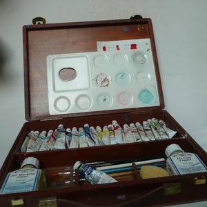 Puede incluir: Caja de pintura de artista de madera abierta, que revela tubos de pintura, una paleta con pintura seca, pinceles y botellas de suministros de arte. La caja es marrón con un asa y cierres de latón.