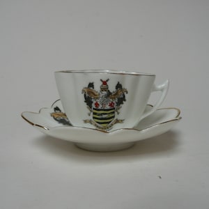 May include: A white teacup and saucer with a gold trim. The cup and saucer have a scalloped edge. The cup has a coat of arms with a lion, a bird, and a shield with a red and white stripe pattern. The saucer has a similar coat of arms but it is smaller.