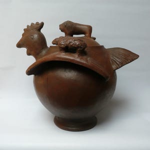Puede incluir: Escultura de cerámica marrón de un recipiente con forma de ave con cabeza de gallo, cola de pez y un pequeño león y oveja en la parte superior. El recipiente tiene un cuerpo redondo y una base de pedestal. La escultura es probablemente hecha a mano.