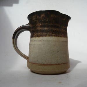 Puede incluir: Una jarra de cerámica con un diseño de esmalte de dos tonos. La parte superior y el asa tienen un acabado en marrón oscuro moteado, mientras que el cuerpo inferior es de color blanco roto con motas. La base es de color tostado con una costura.