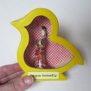 May include: A yellow bird-shaped box with a red and white patterned interior. The box contains a small doll wearing a red and white kimono. The box has a label that reads "Madame Butterfly".