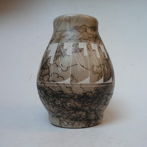 Op de afbeelding: Een beige keramische vaas met een rond lichaam en een smalle hals, voorzien van een donkerzwart, paardenhaarachtig marmerpatroon. Het midden van de vaas is versierd met een horizontale band van witte, gegraveerde geometrische en ovale motieven.
