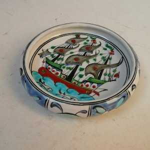 May include: A round, ceramic decorative bowl with a hand-painted design. The bowl features a detailed illustration of a sailing ship with red, green, and grey accents. The rim is decorated with blue and white floral patterns.