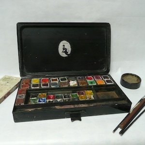 Pode incluir: Um conjunto de tinta de aquarela vintage de metal preto com uma variedade de cores em pequenas bandejas. O conjunto inclui uma tampa com um logotipo que diz "DRUICK'S" e um pequeno recipiente redondo. Há também três pincéis.