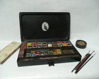May include: A vintage black metal watercolor paint set with a variety of colors in small pans. The set includes a lid with a logo that says "DRUICK'S" and a small round container. There are also three paintbrushes.