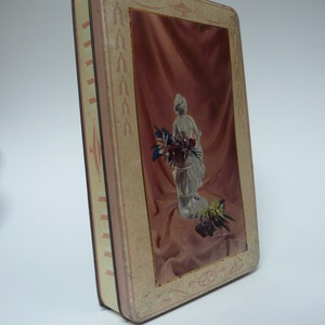 Può includere: Una scatola di latta vintage con un design rosa e bianco. La scatola presenta un'immagine di una donna che tiene un mazzo di fiori. La scatola ha un bordo decorativo con un motivo ripetitivo.