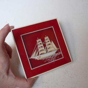 Peut inclure: Une plaque murale décorative carrée présentant un modèle de voilier tridimensionnel de couleur crème centré sur un fond rouge profond. Le navire possède plusieurs mâts et voiles, et la plaque est encadrée par une fine bordure blanche.