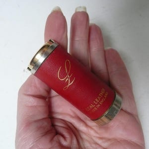 May include: A red leather cylindrical case with gold-coloured metal trim. The case has gold lettering and the words "REAL LEATHER MADE IN ENGLAND".