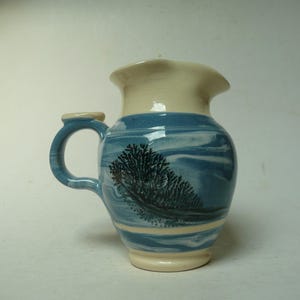 May include: A blue and white ceramic pitcher with a black tree design. The pitcher has a handle and a spout.