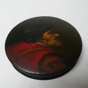 Puede incluir: Un recipiente redondo y negro con un retrato pintado de un hombre con abrigo y sombrero rojos. El retrato es al estilo de una pintura de los viejos maestros, con un fondo oscuro y colores cálidos.