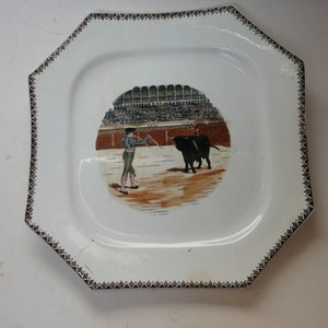 May include: White octagonal ceramic plate with a brown border. The plate features a painted scene of a bullfight in a bullring with a matador and a bull.