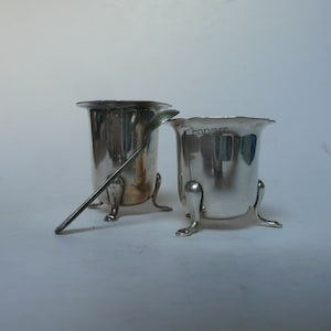 Puede incluir: Dos saleros de plata con una cuchara de plata. Los saleros tienen forma redonda y tres patas. La cuchara está sobre la mesa.