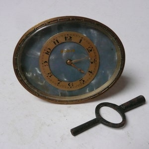 May include: A vintage, oval-shaped, brass alarm clock with a blue and white marbled face. The clock has black Roman numeral hour markers and a key wind mechanism. The clock is accompanied by a black metal winding key.