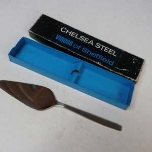 Puede incluir: Un cuchillo para pastel de plata con una caja azul y negra. La caja tiene el texto "Chelsea Steel Viners of Sheffield" impreso en ella.