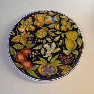Op de afbeelding: Ronde keramische plaat met een donkerblauwe achtergrond en een geschilderd ontwerp van verschillende soorten fruit en groenten. De plaat heeft citroenen, granaatappels, bieten en andere producten, met een witte rand. Het ontwerp is kleurrijk en gedetailleerd.