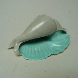 Op de afbeelding: Een keramische zeeschelp met een lichtgrijze buitenkant en een turquoise binnenkant. De opening van de schelp onthult een gedetailleerd, geribbeld patroon in turquoise. De schelp ligt op een lichtgekleurd oppervlak.