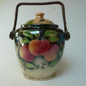 Peut inclure: Un pot à biscuits en céramique vintage avec un couvercle et une anse tressée. Le pot est peint avec un motif de pommes rouges et roses et de feuilles vertes. Le couvercle est beige et vert.