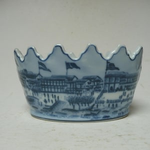 May include: A white ceramic bowl with a scalloped rim, decorated with a blue transferware pattern depicting a landscape scene with buildings, trees, and flags. The design wraps around the exterior of the bowl, featuring detailed architectural structures and figures in a classic blue and white colour scheme.