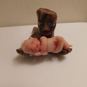 Könnte beinhalten: Eine kleine, braune Tonfigur eines Baumes mit einem Gesicht, die eine schlafende Babypuppe in ihren Armen hält.