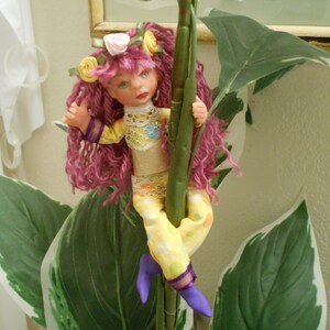 May include: A handmade doll with pink hair, a yellow and white dress, and a flower crown. The doll is holding onto a green stem.