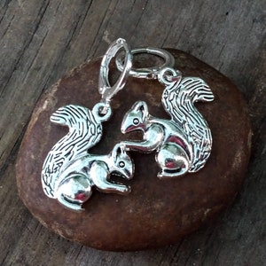 May include: A pair of silver earrings featuring a squirrel design. The squirrels are facing each other and have detailed features. The earrings are attached to silver hoops.