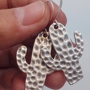 May include: Silver cactus earrings with a hammered texture. The earrings are dangle style with a hook closure.
