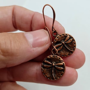 May include: A pair of copper dangle earrings with a dragonfly design. The earrings are made of copper wire and have a round, textured charm with a dragonfly in the center.