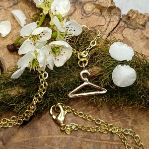 May include: A gold chain necklace with a small gold hanger charm. The necklace is lying on a wooden surface with white flower petals and green moss.