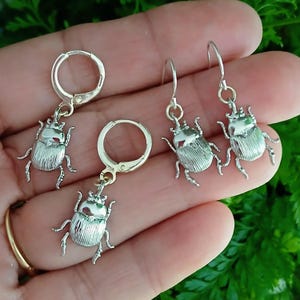 May include: Silver-toned beetle earrings. The earrings feature detailed insect designs with textured wings and delicate legs. One pair has a lever back closure, and the other has a hook closure. The earrings are displayed against a green background.