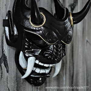Può includere: Maschera da samurai nera e dorata con corna e zanne. La maschera presenta un design dettagliato con intagli intricati e una rete bianca che copre gli occhi.