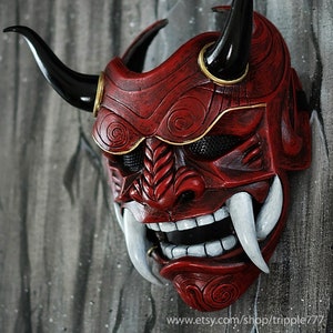 May include: A red and black Oni mask with white fangs and gold accents. The mask has a detailed design with swirling patterns and a mesh eye covering.