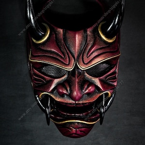 May include: A red and black Oni mask with gold accents. The mask has a fierce expression with sharp teeth and horns. The eyes are covered with a black mesh.