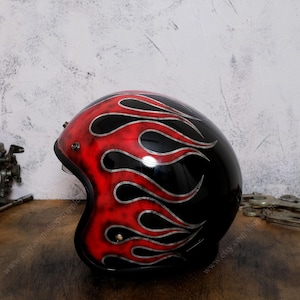 May include: A glossy black motorcycle helmet featuring a prominent red flame design with silver outlines. The helmet has a rounded open-face profile with visible metal hardware accents on the side and a smooth, reflective finish throughout.