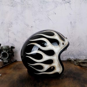 Può includere: Un casco da moto nero lucido caratterizzato da un disegno a fiamme bianche dipinto a mano che si estende lungo il lato. Il casco presenta una calotta liscia e arrotondata con dettagli metallici visibili vicino all'apertura e un profilo aperto.