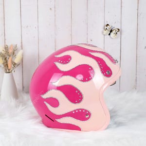 Pode incluir: Um capacete de motocicleta rosa brilhante com padrões de chamas rosa claro delineados com pequenos strass transparentes. O capacete tem um design de casco liso e arredondado com um fecho metálico visível perto da área da orelha.