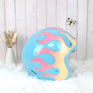 Pode incluir: Um capacete de motocicleta azul-claro com um design de chamas em degradê rosa e amarelo. Os contornos das chamas são delineados com pequenos strass transparentes que refletem a luz. O capacete possui um acabamento liso e brilhante.
