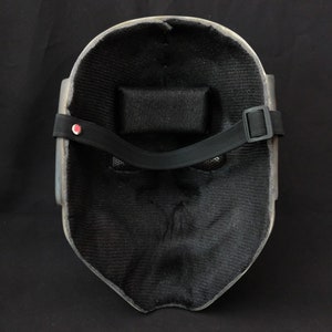 May include: A black and gray mask with a black strap and a mesh covering the eyes. The mask is made of a hard material and has a textured surface.