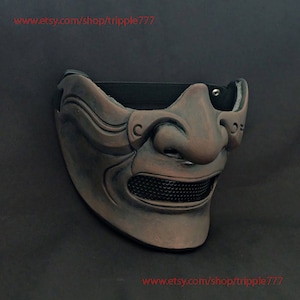Op de afbeelding: Een bruin en zwart samurai-masker met een gaasmondbescherming. Het masker heeft een gestructureerd, gebeeldhouwd ontwerp en is gemaakt van een hard materiaal.