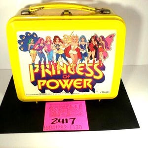 May include: A yellow metal lunchbox featuring a colorful graphic of the Princess of Power characters. The lunchbox has a handle and a clasp closure. The front of the lunchbox has the words "Princess of Power".