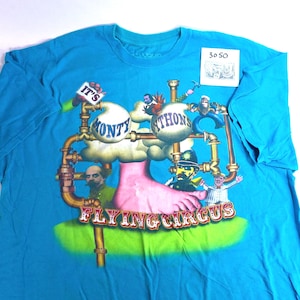 May include: A turquoise blue t-shirt featuring a colorful graphic design. The design includes the text "It's Monty Python's Flying Circus" with cartoon characters and a pink foot. The shirt has a crew neck.