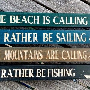 May include: Four wooden signs with different sayings about the beach, sailing, mountains, and fishing. The signs are painted in various shades of blue, green, brown, and black. The text is white and the signs have small icons carved into the wood.