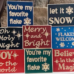May include: Set of nine wooden signs with gold lettering and snowflake designs. The signs feature various Christmas-themed phrases, including "You're my favorite flake", "Let it snow", "Oh Holy Night", "Merry and Bright", "Flakes Welcome", "Joy to the world", "You're my favorite flake", and "Believe in the Magic".