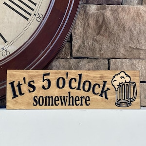 May include: Wooden sign with the phrase "It's 5 o'clock somewhere" in black lettering, accompanied by a beer mug illustration. The sign is made of light-colored wood and is set against a stone wall and a clock.