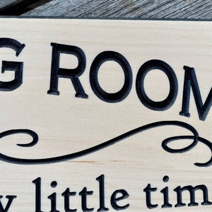 May include: A wooden sign with black lettering that reads "Living Room - a little time". The sign has a decorative flourish between the two lines of text.