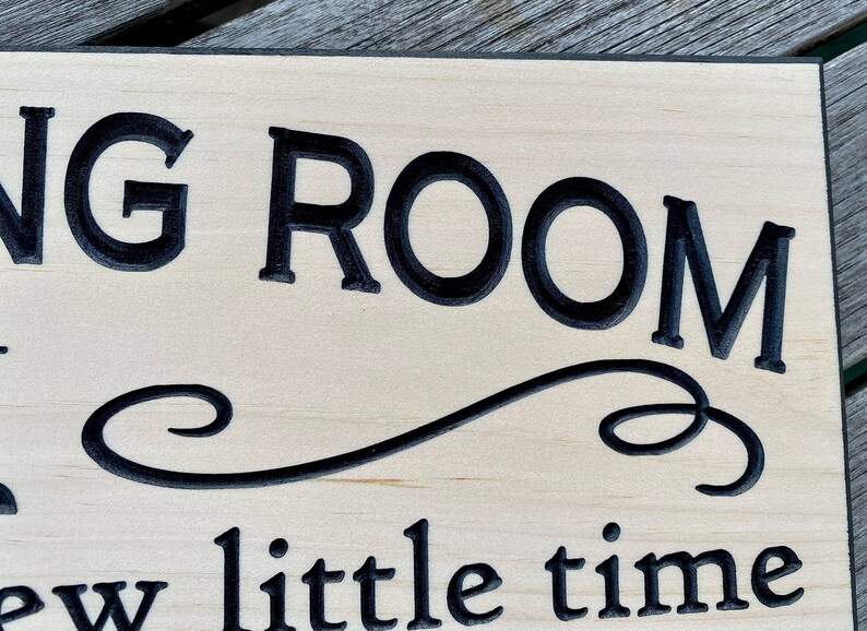 May include: A wooden sign with black lettering that reads "Living Room - a little time". The sign has a decorative flourish between the two lines of text.